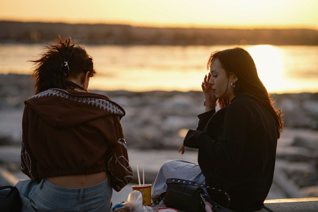 Two women sitting side by side by the water at sunset, together yet each at ease